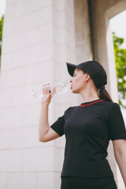 Genç atletik güzel esmer kız siyah üniforma ve kap holding şişe, içme suyu ayakta açık havada şehir parkında çalıştırmadan önce eğitim sırasında. Fitness, sağlıklı yaşam konsepti