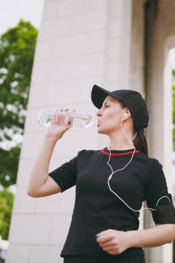 Siyah üniformalı genç atletik güzel esmer kız kap kulaklıklarla müzik ayakta Şehir Park açık havada dinleme eğitimi sırasında şişe içme suyu tutarak. Fitness, sağlıklı yaşam tarzı