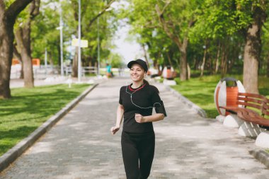 Atletik esmer kız siyah üniforma giyen ve kulaklık ile kap gülümseyen genç spor yapıyor, çalıştıran, koşu, city Park açık havada yolda müzik eğitimi. Fitness, sağlıklı yaşam konsepti