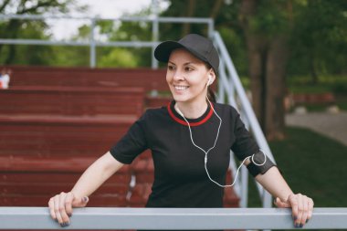 Genç kız siyah üniforma giyen ve kap dinleme müzik, dinlenme ve ayakta tutma önce veya sonra şehir Park açık havada çalışan, eğitim kulaklık ile gülümseyen atletik. Fitness, sağlıklı yaşam konsepti