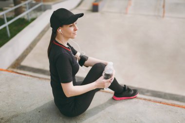 Siyah üniformalı genç atletik gülümseyen kız kap önce veya sonra şehir Park açık havada çalışan, eğitim oturan su ile şişe tutan müzik dinlerken kulaklık ile. Fitness, sağlıklı yaşam tarzı