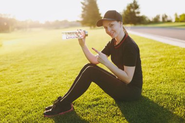 Siyah üniformalı genç kız kap rahatlatıcı yeşil çimenlerin üzerine çalışan, spor egzersizleri sonra içme suyu şişe açık havada güneşli yaz gününde üzerinde dinlenme oturuyor. Fitness, sağlıklı yaşam konsepti