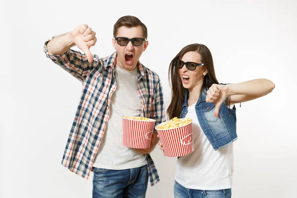 Young irritated couple, woman and man in 3d glasses and casual clothes watching movie film on date, holding buckets of popcorn, showing thumbs down isolated on white background. Emotions in cinema