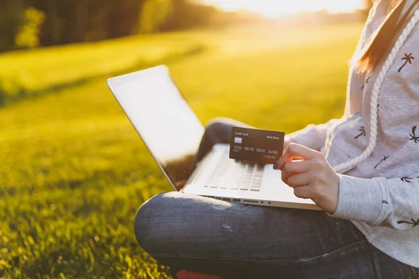 Young happy student female with credit card. Woman sitting on grass ground, working on laptop pc computer in city park on green grass sunshine lawn outdoors. Mobile Office. Freelance business concept