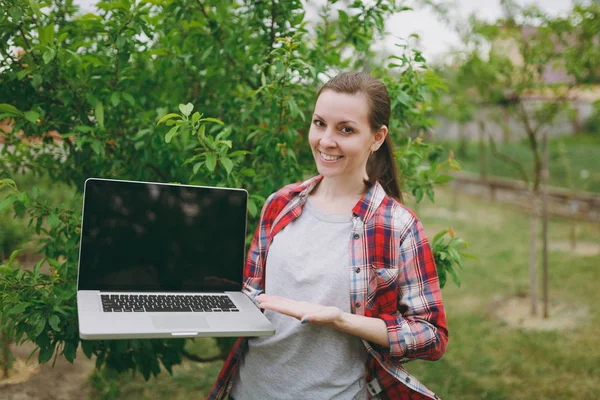 Young pretty woman in checkered shirt standing in garden near green tree, holding modern pc computer laptop with blank black empty screen. Copy space for advertisement. With place for text or photo