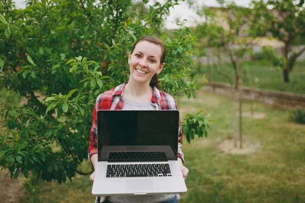 Young pretty woman in checkered shirt standing in garden near green tree, holding modern pc computer laptop with blank black empty screen. Copy space for advertisement. With place for text or photo