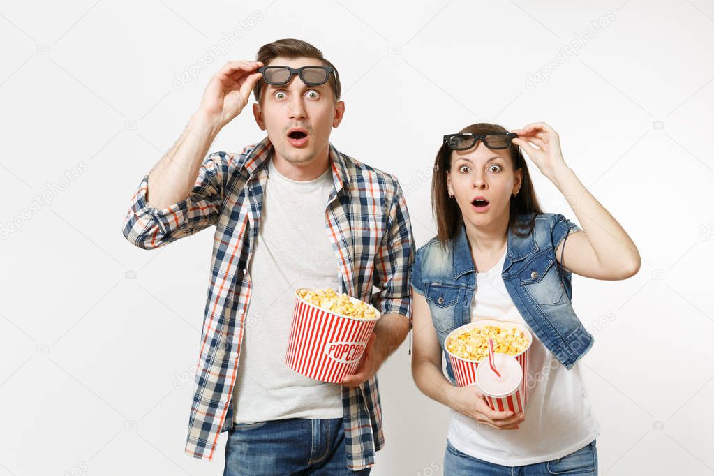 Young shocked couple, woman and man watching movie film on date, holding bucket of popcorn, plastic cup of soda or cola, taking off 3d glasses isolated on white background. Emotions in cinema concept