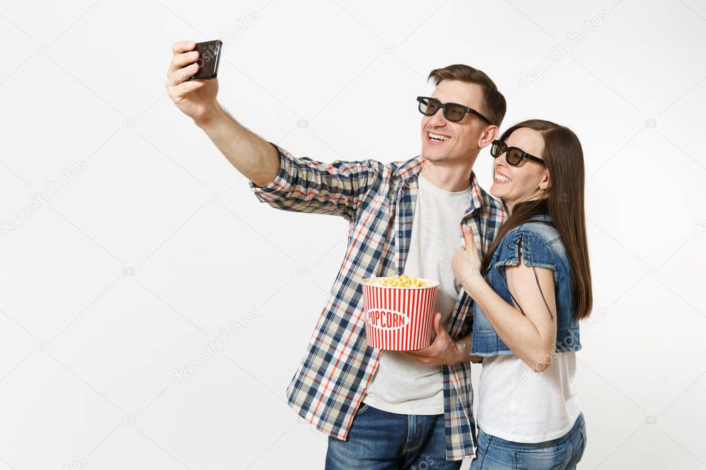 Young couple, woman and man in 3d glasses watching movie film on date, holding bucket of popcorn and cup of soda or cola, doing selfie on mobile phone isolated on white background. Emotions in cinema