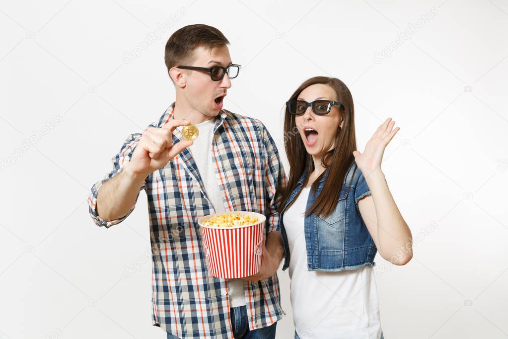 Young shocked couple, woman and man in 3d glasses and casual clothes watching movie film on date, holding bucket of popcorn and bitcoin isolated on white background. Emotions in cinema concept