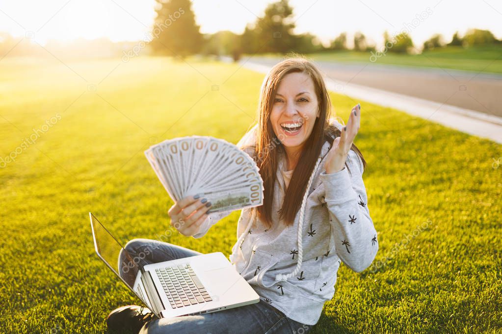 Female holding bundle of dollars cash money. Woman sitting on grass ground, working on laptop pc computer in city park on green grass sunshine lawn outdoors. Mobile Office. Freelance business concept