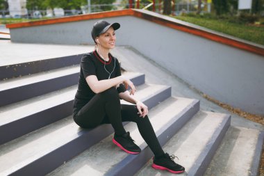 Genç atletik gülümseyen kız siyah üniforma ve dinlenme ve önce veya sonra çalışan, eğitim şehir park açık havada merdivenlerde oturan müzik kulaklık ile kap. Fitness, sağlıklı yaşam tarzı