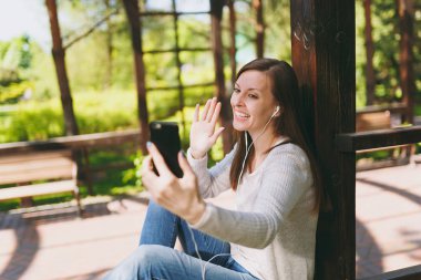 Hafif rahat kıyafetler giymiş güzel kadın portresi. Şehir Parkı açık havada bahar doğa üzerinde sokakta oturan kadın gülümsüyor, selfie yaparken cep telefonu veya görüntülü arama ateş etti. Yaşam tarzı kavramı