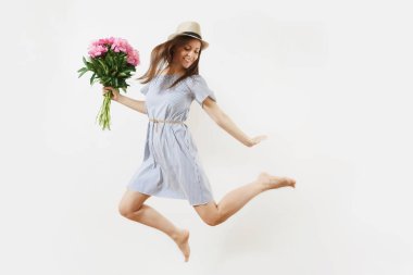 Beyaz arka plan üzerinde atlama genç kadın mavi elbiseli, buket pembe peonies çiçek, holding şapkası izole mutlu eğlenceli uzunluğu tam. Sevgililer günü, Dünya Kadınlar günü tatil kavramı