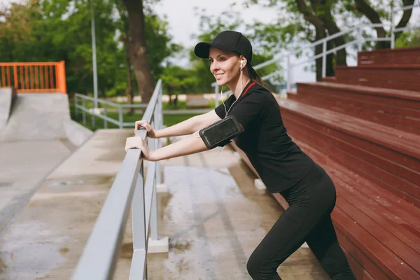 Atletik güzel esmer kız siyah üniforma, gülümseyen genç kap kulaklıklarla müzik ısınma-up şehir park havada spor germe egzersizleri yapıyor. Fitness, sağlıklı yaşam tarzı