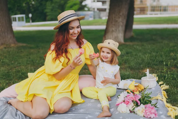 Smiling woman in yellow clothes rest, eat macaroons and showing dessert on camera with little child baby girl in park. Mother, little kid daughter. Mother\'s Day, love family, parenthood, childhood