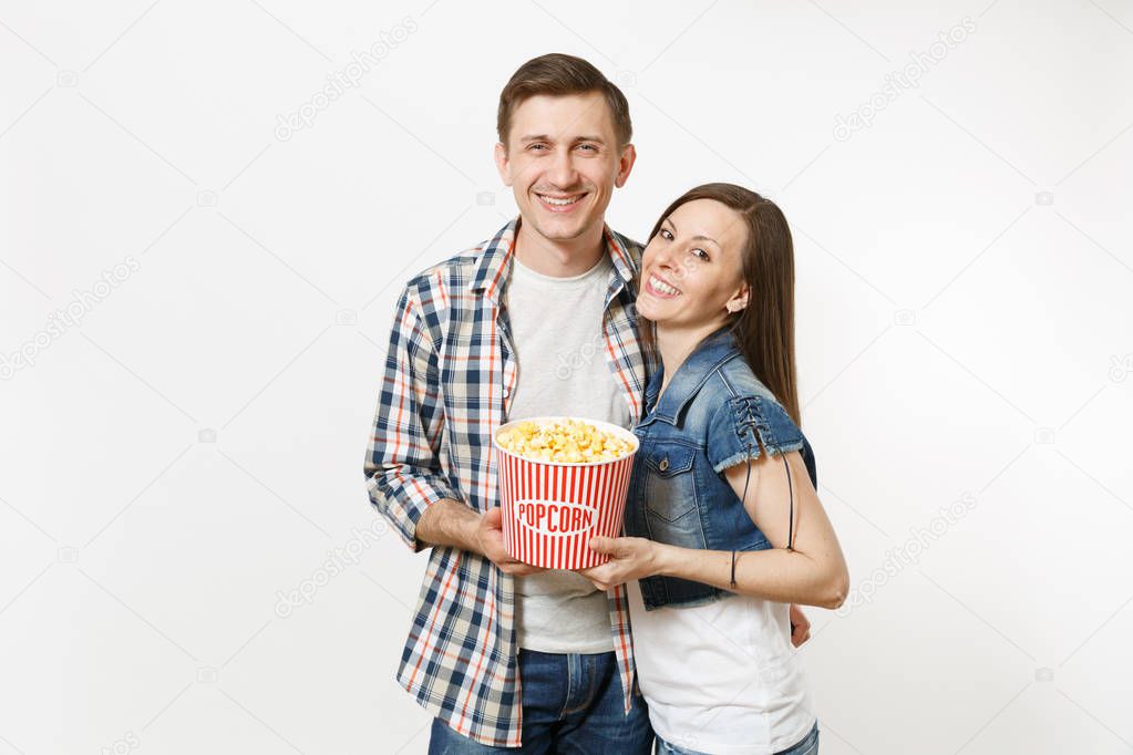 Young happy smiling couple, woman and man in casual clothes watching movie film on date, holding bucket of popcorn, hugging isolated on white background. Emotions in cinema concept