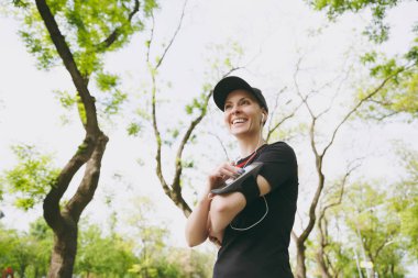 Kulaklıklarla müzik, siyah üniformalı genç gülümseyen atletik kız uygulaması çalıştıran veya üstünde hareket eden telefon, koşu için app kullanarak şehir parkı mekanlarda eğitim. Fitness, sağlıklı yaşam tarzı