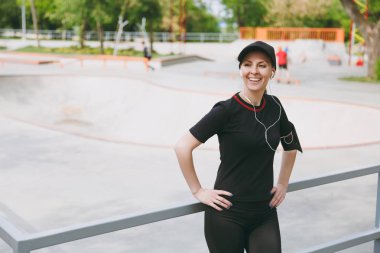 Açık havada şehir parkında eğitim genç atletik gülümseyen esmer kız siyah üniforma ve müzik, önce veya sonra çalışan, ayakta dinlerken kulaklık ile kap. Fitness, sağlıklı yaşam konsepti