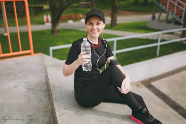 Siyah üniformalı genç atletik gülümseyen kız kap önce veya sonra şehir Park açık havada çalışan, eğitim oturan su ile şişe tutan müzik dinlerken kulaklık ile. Fitness, sağlıklı yaşam tarzı