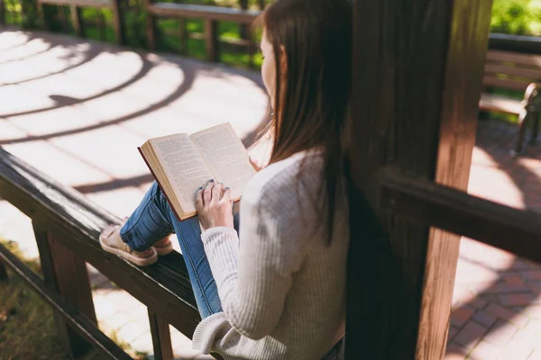 Portrait of calm peaceful young woman wearing light casual clothes relaxing, reading book. Female resting in city park in street outdoors on spring nature. Lifestyle concept. Back view. Copy space