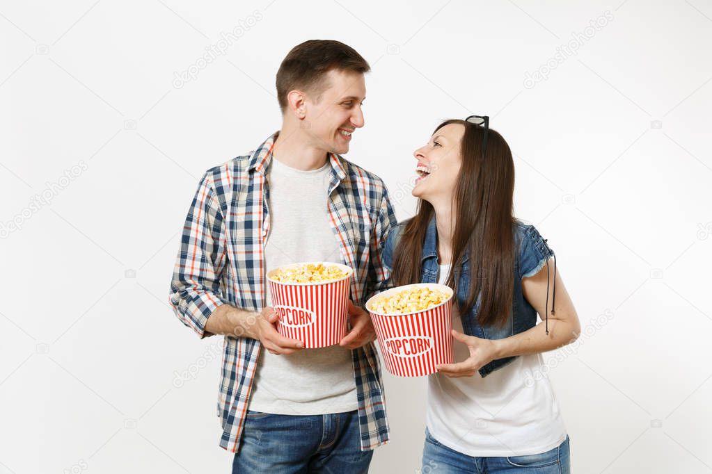 Young happy smiling couple, woman and man in 3d glasses and casual clothes watching movie film on date, holding buckets of popcorn, laughing isolated on white background. Emotions in cinema concept