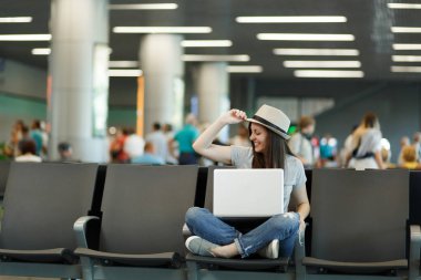 Genç gülümseyen şapka, düzleştirme seyahat etmek turist kadın çapraz ayaklı dizüstü bilgisayar ile Uluslararası Havaalanı'nda lobi salonunda bekleyin. Hafta sonu kaçamağı yurt dışında seyahat yolcu. Hava uçuş kavramı