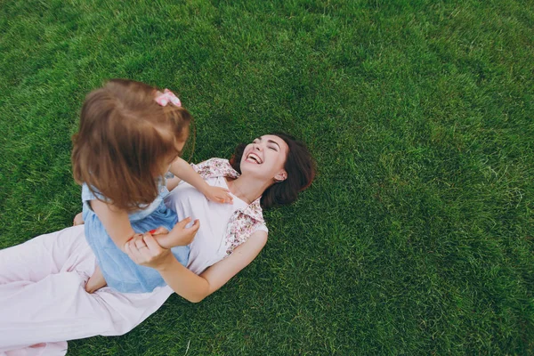 Laughing woman in light dress and little cute child baby girl lie on green grass in park rest, play and have fun. Mother, little kid daughter. Mother\'s Day, love family, parenthood, childhood concept