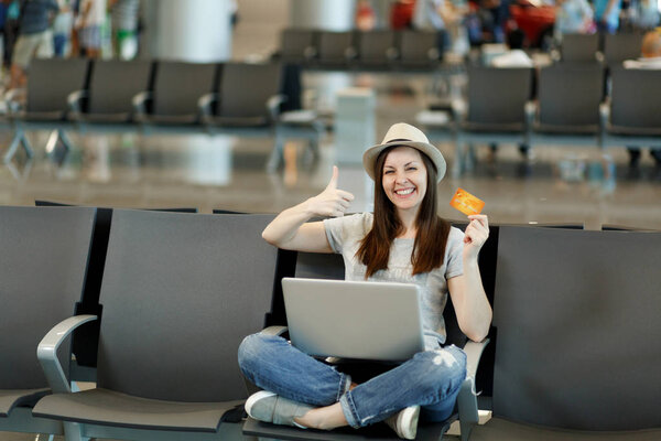 Радостная туристка-путешественница с ноутбуком, сидящая со скрещенными ногами, показывающая большой палец вверх, держащую кредитную карту, ждет в вестибюле аэропорта. Пассажир, выезжающий за границу в выходные. Концепция полета
