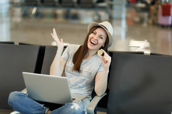Молодая смеющаяся туристка-путешественница в шляпе сидит, работает на ноутбуке, держит биткойн, расправляет руки, ждет в холле аэропорта. Пассажир, выезжающий за границу в выходные. Концепция полета

