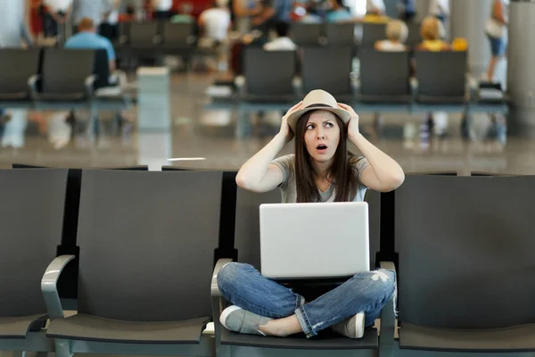 Young concerned traveler tourist woman with laptop sitting with crossed legs clinging to head while waiting in lobby hall at airport. Passenger traveling abroad on weekend getaway. Air flight concept