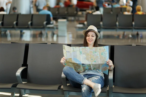 Young pretty traveler tourist woman holding paper map, search route waiting in lobby hall at international airport. Passenger traveling abroad on weekends getaway. Air travel, flight journey concept
