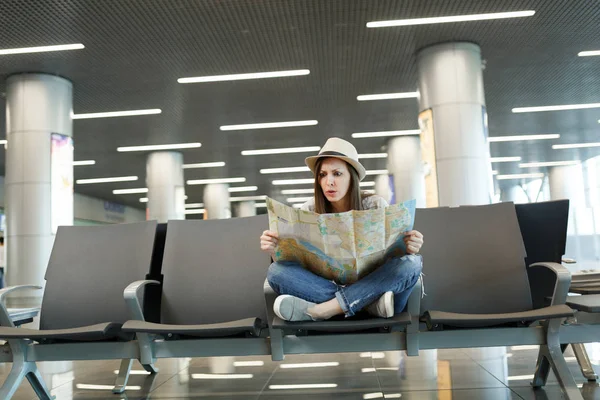 Young scared traveler tourist woman with crossed legs holding paper map, search route waiting in lobby hall at international airport. Passenger traveling abroad on weekend getaway. Air flight concept