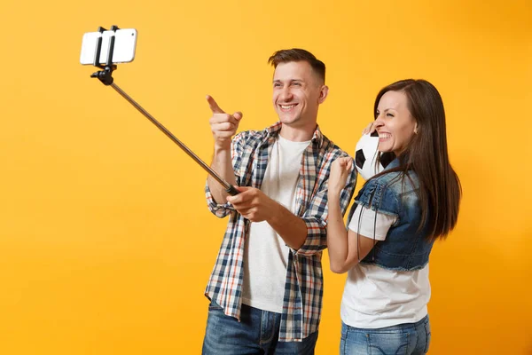 Young couple woman man, football fans doing selfie on mobile phone with monopod selfish stick, cheer up support team, soccer ball isolated on yellow background. Sport family leisure lifestyle concept