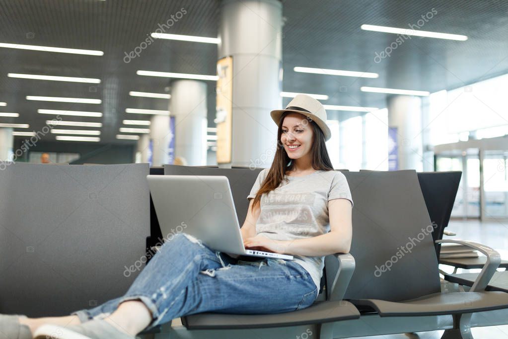 Young pretty traveler tourist woman in hat working on laptop while waiting in lobby hall at international airport. Passenger traveling abroad on weekends getaway. Air travel, flight journey concept