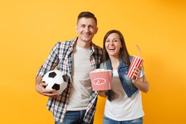 Genç eğlenceli çift, kadın erkek, holding futbol topu, kova patlamış mısır, plastik bardak kola, futbol taraftarları destek ekibi, sarı zemin üzerine izole neşelen. Spor aile eğlence yaşam tarzı kavramı