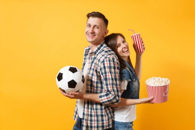 Genç eğlenceli çift, kadın erkek, holding futbol topu, kova patlamış mısır, plastik bardak kola, futbol taraftarları destek ekibi, sarı zemin üzerine izole neşelen. Spor aile eğlence yaşam tarzı kavramı