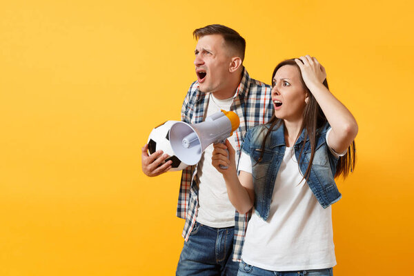 Angry expessive crazy couple, woman man football fans screaming, upset of loss, goal of favorite team with soccer ball, megaphone isolated on yellow background. Sport family leisure lifestyle concept