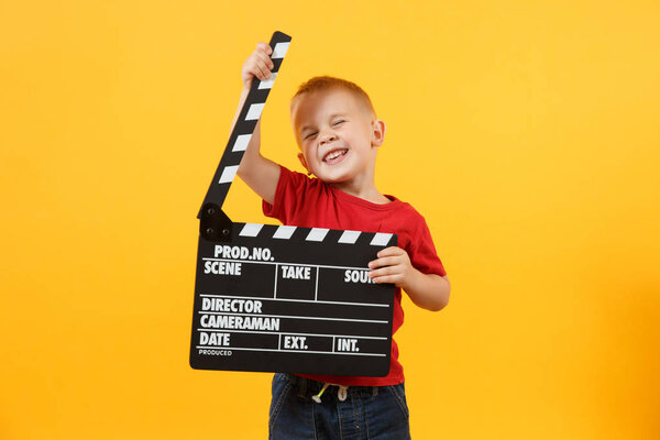 Little cute kid baby boy 3-4 years old in red t-shirt holding in hand classik black film making clapperboard looking camera isolated on yellow background. Kids childhood lifestyle concept. Copy space