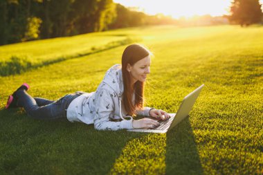 Genç öğrenci rahat kıyafetler içinde kadın akıllı. Çim, dizüstü pc bilgisayar Şehir Park açık havada Güneş çim yeşil çim üzerinde üzerinde çalışıyor yatan kadın. Mobil ofis. Serbest iş kavramı