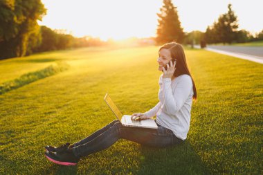Genç öğrenci rahat kıyafetler içinde kadın akıllı. Dizüstü pc bilgisayar Şehir Park açık havada Güneş çim çimen üzerinde çalıştığı kadın üstünde hareket eden telefon, söz. Mobil ofis. Serbest iş kavramı