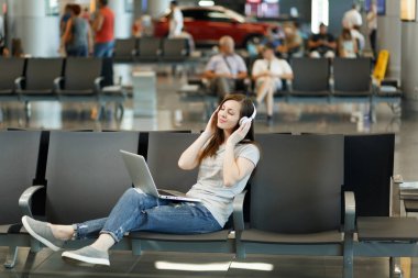 Dizüstü bilgisayarda çalışan müzik dinlerken kulaklık ile seyahat etmek turist kadın genç rahat, Uluslararası Havaalanı'nda lobi salonunda bekleyin. Yurt dışında hafta sonu kaçış üzerinde seyahat yolcu. Hava uçuş kavramı