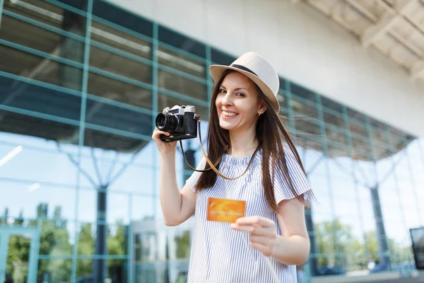 Young happy traveler tourist woman in hat holding retro vintage photo camera, credit card at international airport. Passenger traveling abroad on weekends getaway. Air travel, flight journey concept