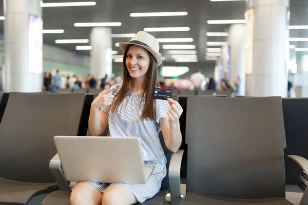 Young traveler tourist woman working on laptop, pointing index finger on credit card, waiting in lobby hall at international airport. Passenger traveling abroad on weekend getaway. Air flight concept