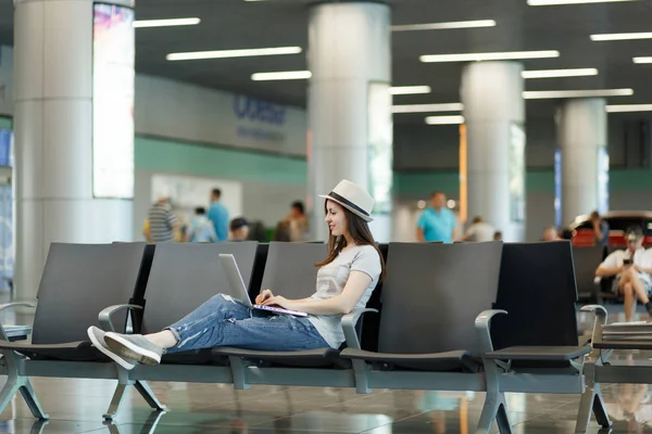 Young calm traveler tourist woman sitting, working on laptop while waiting in lobby hall at international airport. Passenger traveling abroad on weekends getaway. Air travel, flight journey concept