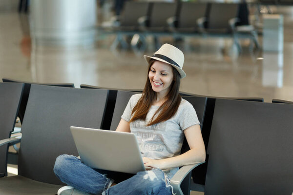 Молодая симпатичная туристка-путешественница в шляпе, сидящая со скрещенными ногами и работающая на ноутбуке, ждет в вестибюле международного аэропорта. Пассажир, путешествующий за границу по выходным. Концепция полета
