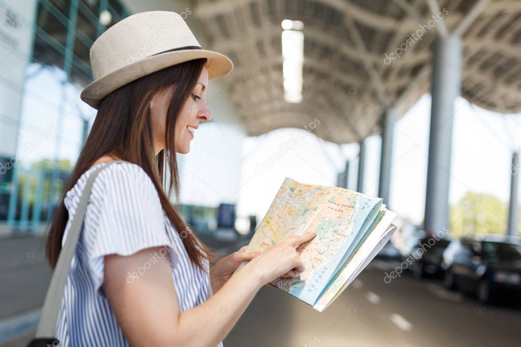 Young smiling traveler tourist woman in hat with backpack searching route in paper map at international airport. Female passenger traveling abroad to travel on weekends getaway. Air flight concept