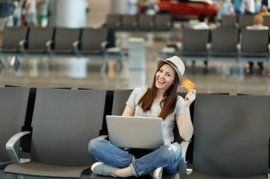 Genç gülümseyen seyahat etmek turist kadın şapkalı çapraz ayaklı otur, dizüstü bilgisayar üzerinde çalışan Havaalanı'nda lobi salonunda kredi kartı bir dakika tutun. Yurt dışında hafta sonu kaçış üzerinde seyahat yolcu. Hava uçuş kavramı