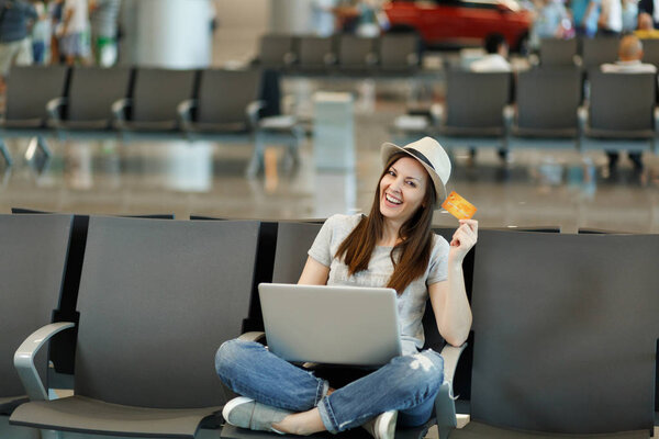 Молодая улыбчивая туристка-путешественница в шляпе сидит со скрещенными ногами, работая на ноутбуке, держит кредитную карту в холле аэропорта. Пассажир, путешествующий за границу по выходным. Концепция полета
