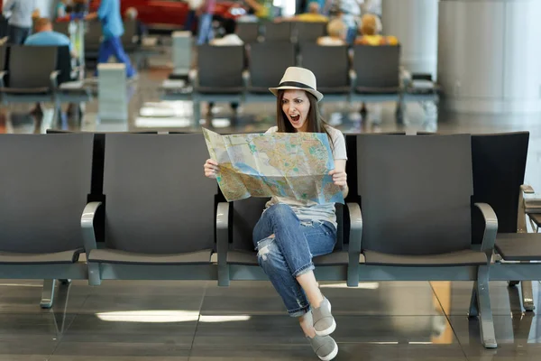 Young irritated traveler tourist woman holding paper map, search route, screaming waiting in lobby hall at airport. Passenger traveling abroad on weekends getaway. Air travel, flight journey concept