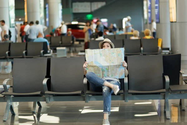 Young traveler tourist woman covering with paper map, search route, waiting in lobby hall at international airport. Passenger traveling abroad on weekends getaway. Air travel, flight journey concept